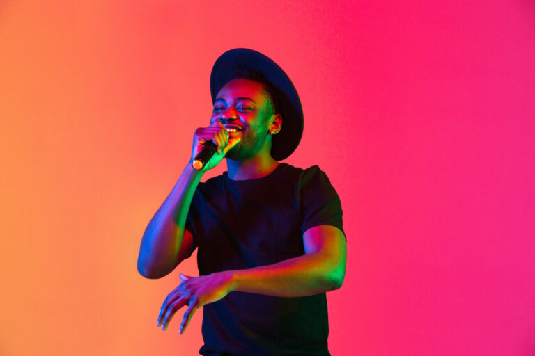 Young african-american musician singing on gradient orange-purple studio background in neon light. Concept of music, hobby, festival. Joyful party host, stand upper. Colorful portrait of artist.