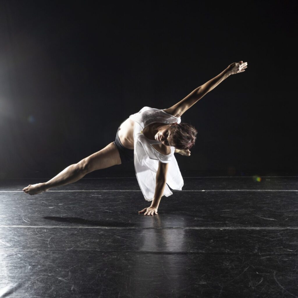 Dancer Angela Gallo performs a floor trick on stage, with her body low to the ground and one leg extended.