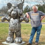 Bob Doster, sculptor, posing with a metal life-sized art piece of a bear made of many hand pieces.