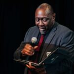 Arts Administrator Kyle C. Coleman presenting on a stage with a microphone.